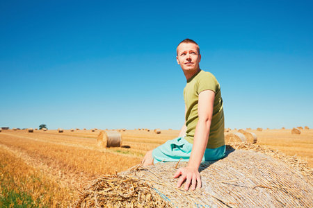 Cornfield after harvest. Young man (farmer) sitting on the bale of straw on the field.の写真素材