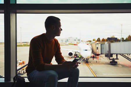 Silhouette of the young man with mobile phone and boarding pass in hand waiting inside airport terminal. The ground crew prepares the plane for the next flight.の写真素材