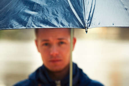 Rainy day. Young man is holding blue umbrella and walking in rain. - selective focus on the dropの写真素材