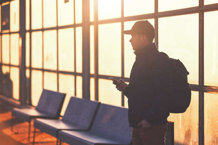 Young traveler with mobile phone in hand waiting at the bus station during sunset.の写真素材