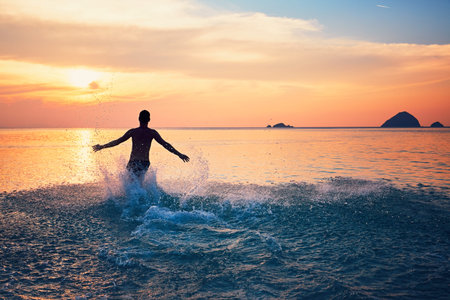 Joyful young man running in water of the tropical sea during beautiful sunset.