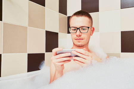Young man sitting in the bathtub and using smart phone to watch tv (video) or texting. Themes for connection, social networking and modern addiction.の写真素材