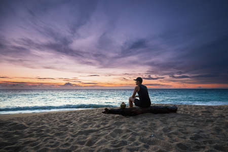 Pensive young man with coconut drink relaxing on the beach during beautiful sunset.の写真素材