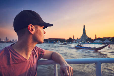 Young man (tourist) on the boat watching the traditional boats against temple Wat Arun. Bangkok at the sunset, Thailand.の写真素材