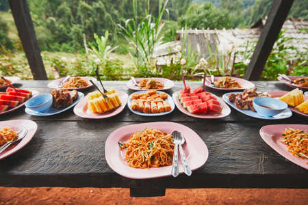 Lunch in the middle of the jungle. The table full of foods and fruits. Traditioal Pad Thai, spring rolls, melon and pineapple. Chiang Mai Province, Thailand.の写真素材