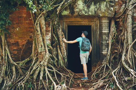 Young man with backpack coming to ancient monuments under the giant roots of the tree near Siem Reap (Angkor Wat) in Cambodiaの写真素材