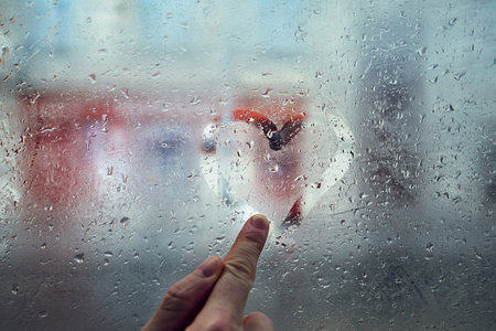 Finger of the young man drawing heart shape on the window with raindrops against city street.
