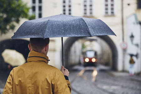 Young man holding umbrella walking in rain. Old city street in Prague, Czech Republicの写真素材