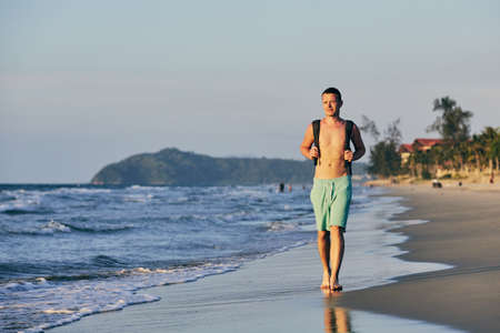 Young man (tourist) with backpack walking on sand beach.の写真素材