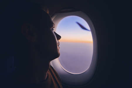 Silhouette of young man traveling by airplane. Pensive passenger looking through window during flight at sunrise.の写真素材