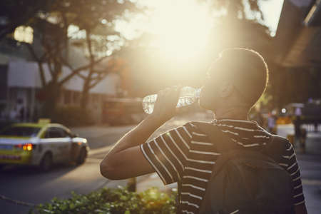 Young man drinking water from bottle during walk on street at sunset.の写真素材