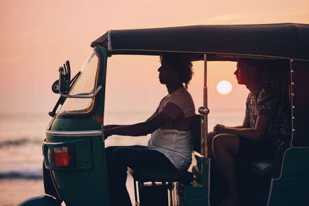 Driver and passenger are traveling by tuk tuk taxi against sea at sunset in Sri Lanka.の写真素材