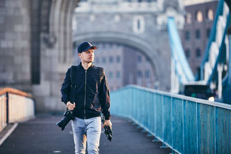 Photographer looking away while carrying camera and tripod against Tower Bridge. London, United Kingdom.の写真素材