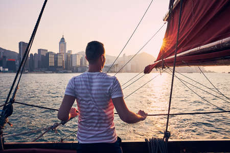 Traveler on traditional junk boat crossing Victoria Harbor against Hong Kong cityscape with skyscrapers at golden sunset.の写真素材