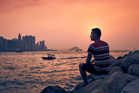 Young man sitting on stones against Hong Kong cityscape with skyscrapers at golden sunset.の写真素材