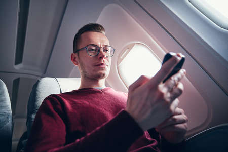 Internet connection during flight. Young man with eyeglasses using mobile phone in airplane.の写真素材