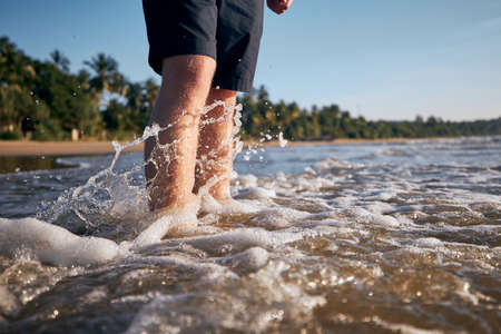 Legs of young man standing in sea against tropical sand beach.の写真素材
