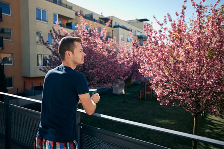 Spring morning at home. Young man with cup tea or coffee on balcony.