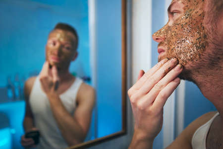 Young man applying facial mask in front of mirror in home bathroom. Themes skin care, treatment and mens beauty.の写真素材