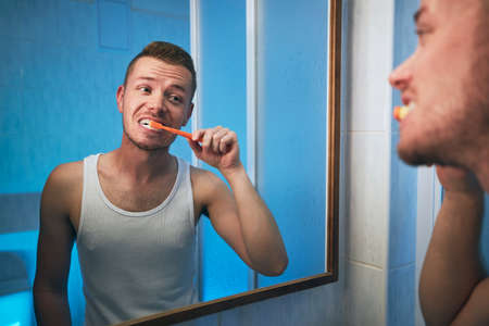 Morning hygiene routine. Man brushing teeth and looking in mirror of home bathroom.の写真素材