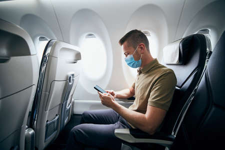 Man wearing face mask and using phone inside airplane during flight. Themes new normal, coronavirus and personal protection.の写真素材