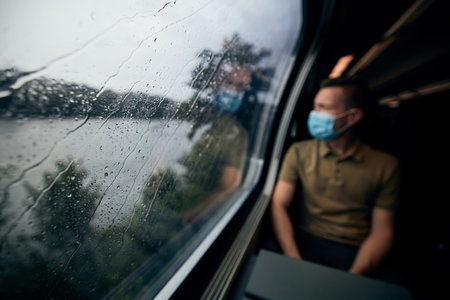 Man wearing face mask inside train in rainy day. Selective focus on raindrops on window.