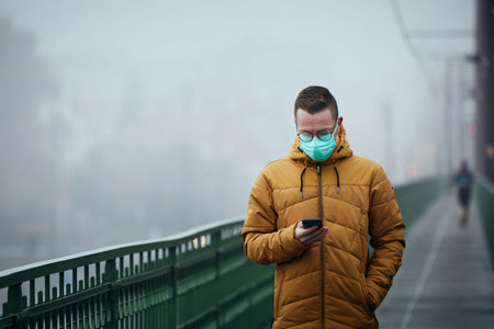 Lonely man with face mask using phone during walk on bridge against city in mysterious fog. Gloomy...