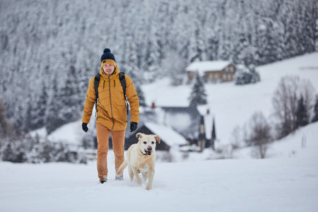 Young man with dog in winter. Pet owner with his labrador retriever walking against old village in beautiful nature. Jizera mountains, Czech Republicの写真素材