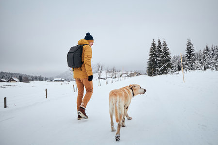 Rear view of young man with dog in winter. Pet owner with his labrador retriever walking against old village in beautiful nature. Jizera mountains, Czech Republicの写真素材