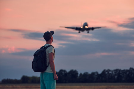 Young man looking up at flying airplane against moody sky during dusk. Themes nostalgia for travel...