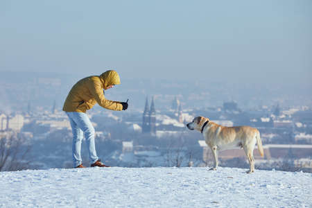 Pet owner taking photo of his labrador retriever via smart phone during frosty day. Young man with dog in winter against urban skyline. Prague, Czech Republicの写真素材