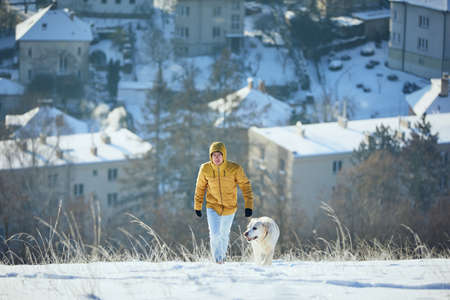 Happy young man with dog in winter. Pet owner with his labrador retriever walking in snow against city. Prague, Czech Republicの写真素材