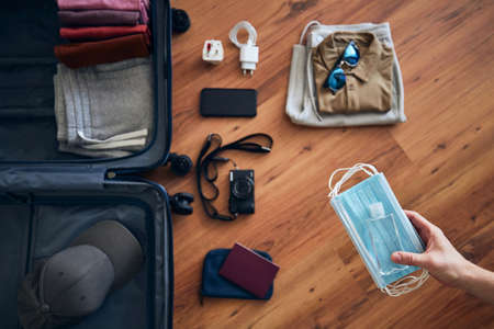 Traveler is preparing for travel in new normal. Hand holding face masks and hand sanitizer for personal protection against packing suitcace with summer clothing, passport and camera.の写真素材