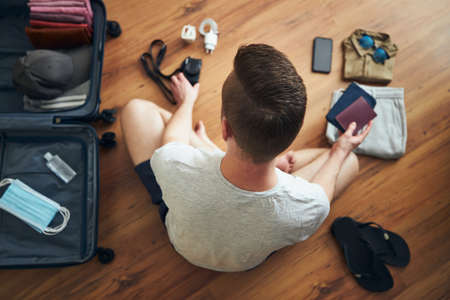 Traveler is preparing for travel in new normal. Man packing suitcase with passport, smart phone, clothing and face masks and hand sanitizer for personal protection.の写真素材