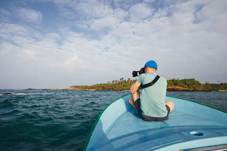 Photographer sitting on boat. Young man photographing beautiful coast with palm tree near Tangalle in Sri Lanka.の写真素材