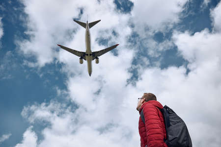 Young man looking up at flying airplane against cloudy sky. Themes nostalgia for travel and aviation.の写真素材