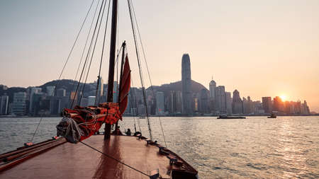 Traditional Junk boat crossing Victoria Harbour against Hong Kong urban skyline with skyscrapers at beautiful sunset.の写真素材