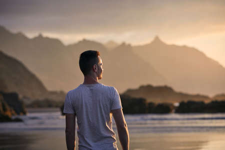 Rear view of pensive man on beach aganinst cliff at golden sunset. Beautiful nature in Tenerife. Canary Islands, Spain.の写真素材