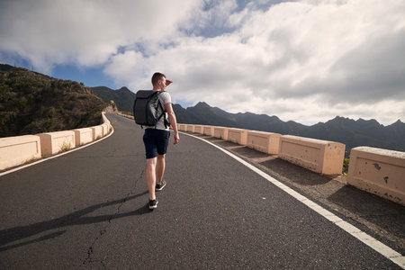 Rear view of man with backpack on mountain road. Young tourist enjoys trip on sunny day. Tenerife, Canary Islands, Spain.の写真素材