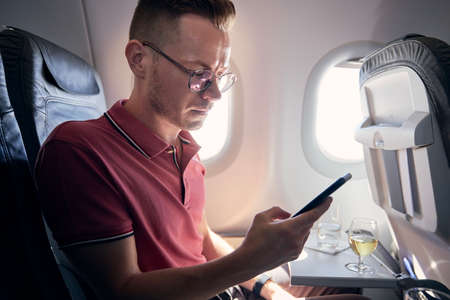 Young man using mobile phone at airplane. Passenger enjoys flight with internet connection a beverages.の写真素材