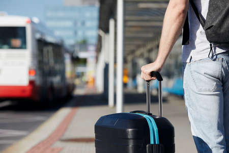 Passenger waiting at bus station in front of airport terminal. Selective focus on hand holding suitcace.の写真素材