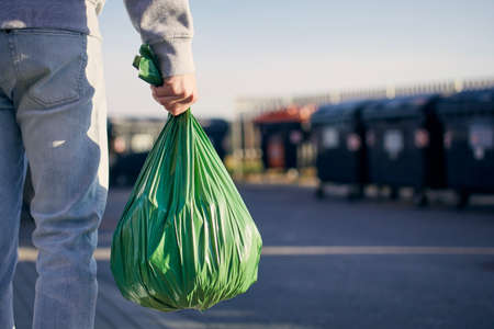 Man walking with rubbish. Rear view of person carrying plastic bag against garbage cans on street.の写真素材