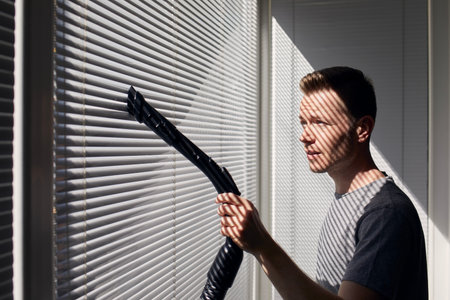 Man cleaning dust from window blind by vacuum cleaner at home. Themes housework and housekeeping.の写真素材