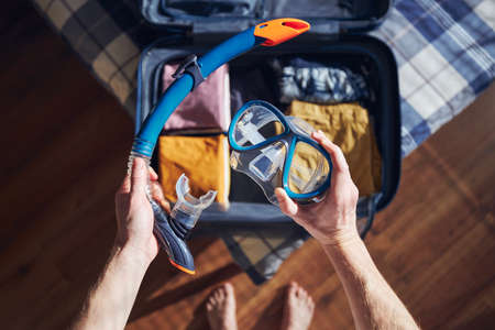 Young man holding snorkel and goggles in hands at home. Packing suitcase and planning travel adventure.の写真素材