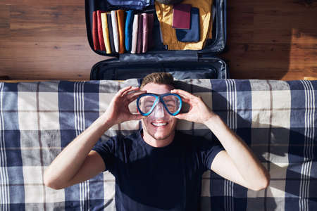 Funny portrait of happy young man during packing suitcase and planning travel adventure.の写真素材