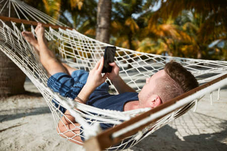Man swinging in hammock and using smart phone. Relaxation on white sand beach under palm trees.の写真素材