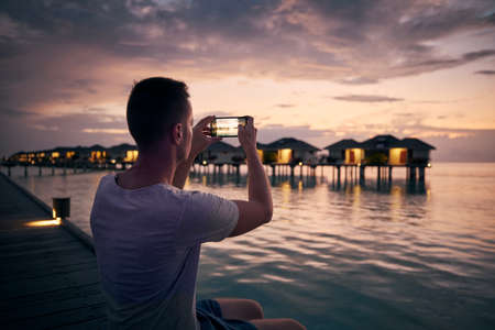 Young man with mobile phone photographing seascape with water bungalows at beautiful sunset in Maldives.の写真素材