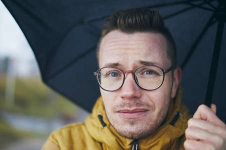 Portrait of young man with eyeglasses under umbrella in rain. Themes weather a meteorology.の写真素材