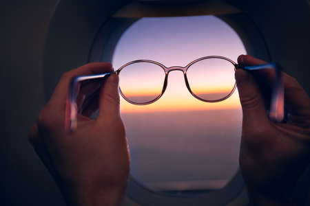 Hand of passenger holding eyeglasses against airplane window during colorful sunset. Themes travel, eyesight and vision.の写真素材