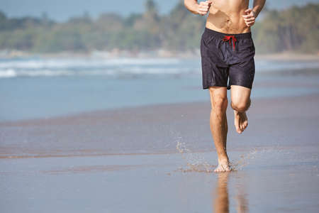 Man running on sand beach. Front view of barefoot runner against coastline.の写真素材